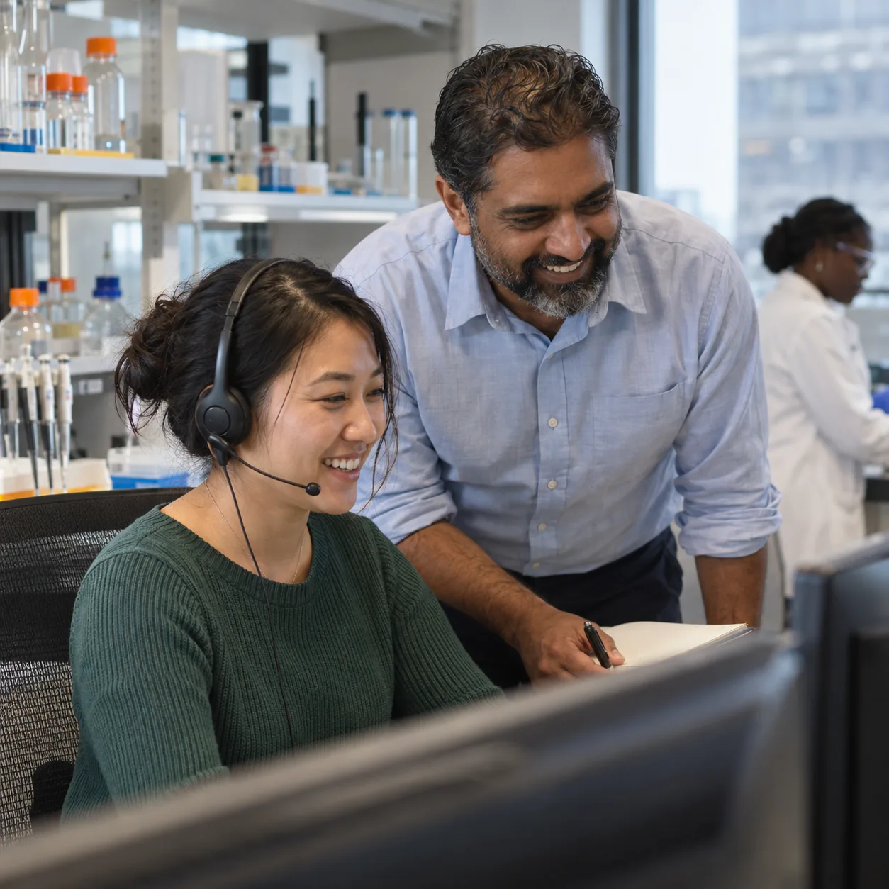 A senior rep coaches a smiling new colleague on a headset in a biotech office